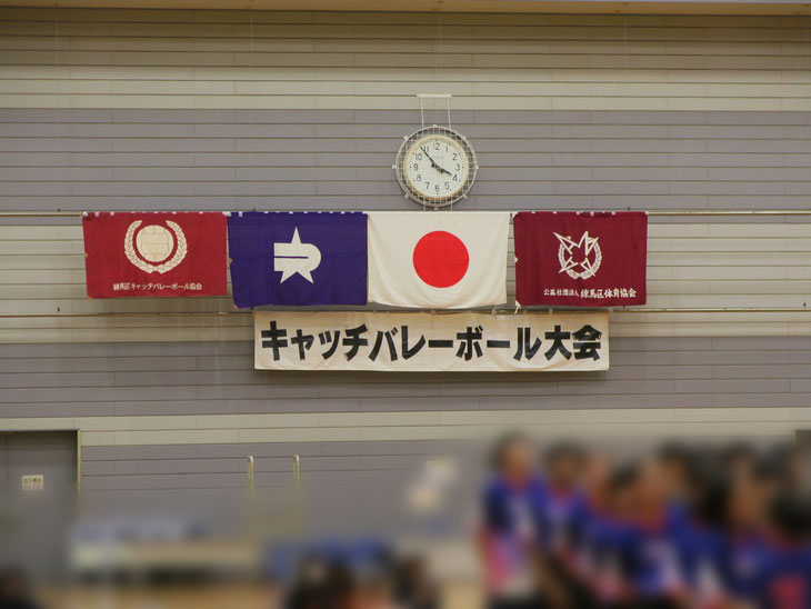 キャッチバレーボール大会の字幕と体育館の壁にかかる旗。左から練馬区キャッチバレー協会、練馬区、国旗、練馬区体育協会。