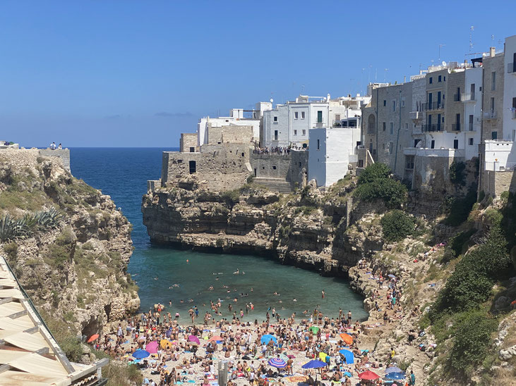 spiaggetta affollata contornata da alti scogli con sopra case bianche e in fondo il mare blu