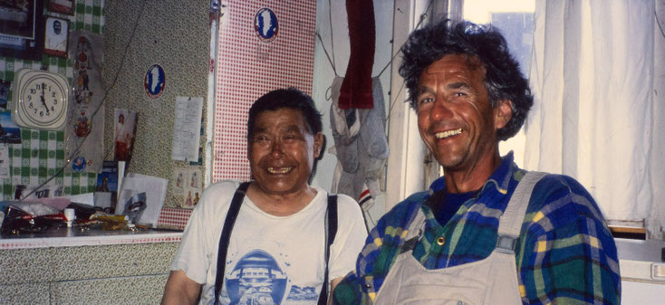 En stue i Østgrønland i 1980'erne. I baggrunden hænger sokker til tørre. Til højre i billedet ses Robert Peroni, der smiler bredt. Til venstre for ham ses en inuit, der også smiler bredt. Hans smil afslører en lang tandmangel i overkæben.