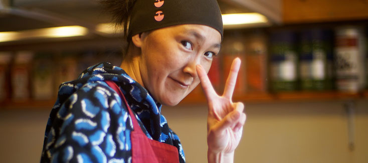 A young Inuit woman looks into the camera and makes the victory sign with her index and middle fingers. She smiles. The picture was taken in the kitchen of The Red House.