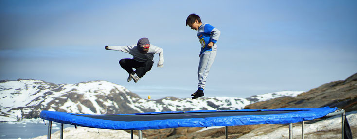 Blauer Himmel, im Hintergrund verschneite Berge. Zwei Inuit-Jungen springen auf einem Trampolin.