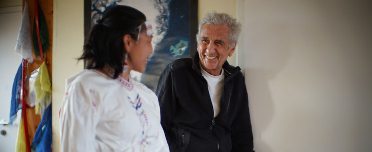 At The Red House Hotel. On the right is Robert Peroni, laughing, around 70 years old. To his left is an Inuit woman in traditional clothing. She is seen from the side. Robert Peroni and the woman are looking at each other.