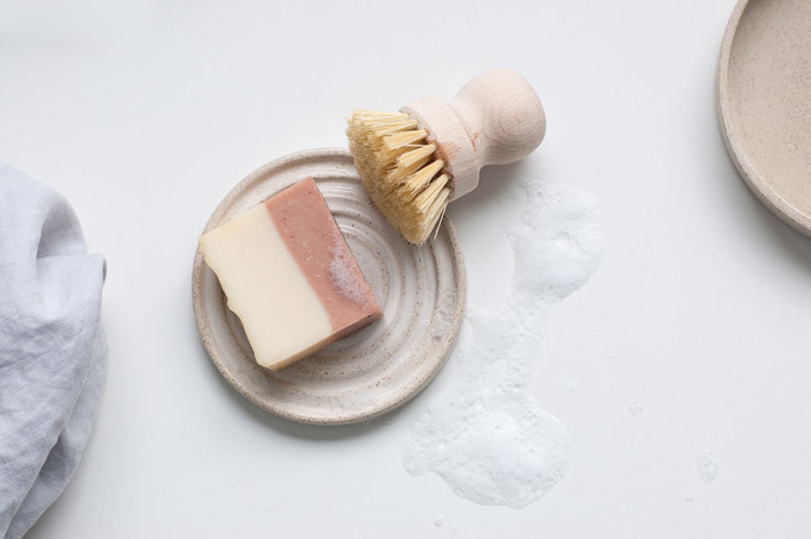 Handmade ceramic soap dish with a bar of soap on top of it and a brush, some foam and a towel next to it