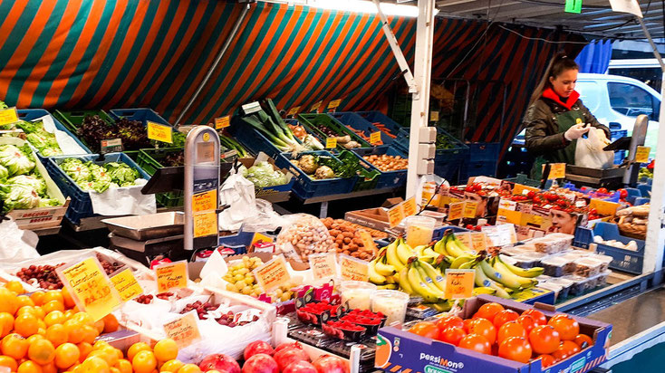 Marktstand mit Obst und Gemüse sowie Verkäuferin, die Ware abwiegt