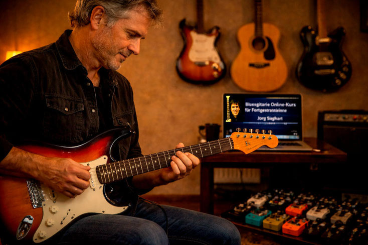 Ein E-Gitarrist (ca. 50 Jahre) spielt eine Stratocaster E-Gitarre, im Hintergrund Gitarren an den Wänden, ein Laptop mit dem Kurs "Bluesgitarre Online-Kurs für Fortgeschrittene" von Jörg Sieghart und am Boden diverse Gitarrenpedale