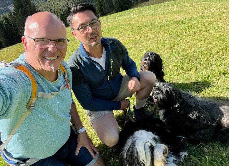 Zwei Personen und vier Tibet Terrier auf einer sommerlichen, saftig grünen Bergwiese