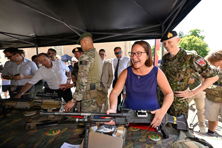 ▲La ministre de la Guerre, Yuriko Backes, dans une robe d’été colorée, arborant un large sourire tout en brandissant un fusil mitrailleur.