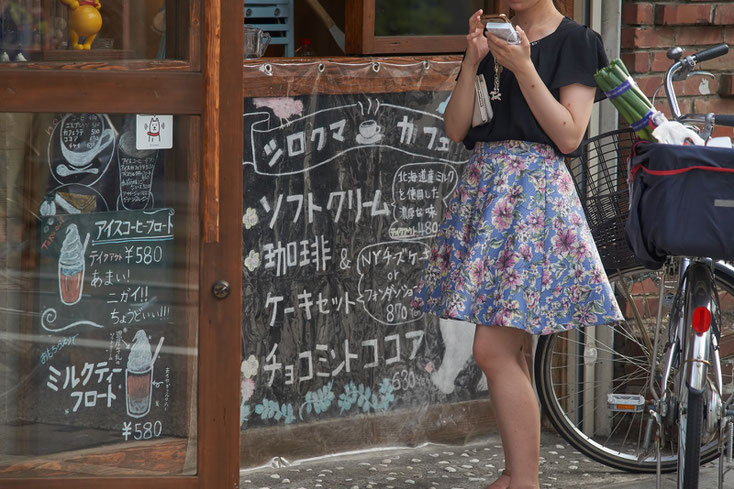 今風のお洒落なCafeの前で、花柄のスカートをはいた女性がスマホをいじっている