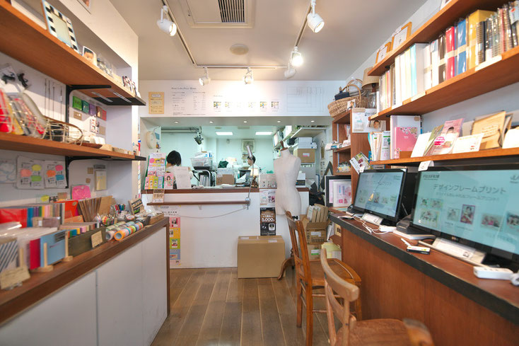 フォトカノンの店内。白い壁に明るい雰囲気。左側に商品(小物)が陳列されている。右側には客が座って作業できるカウンター。突き当り奥のカウンターの向こう側でスタッフが作業をしている。