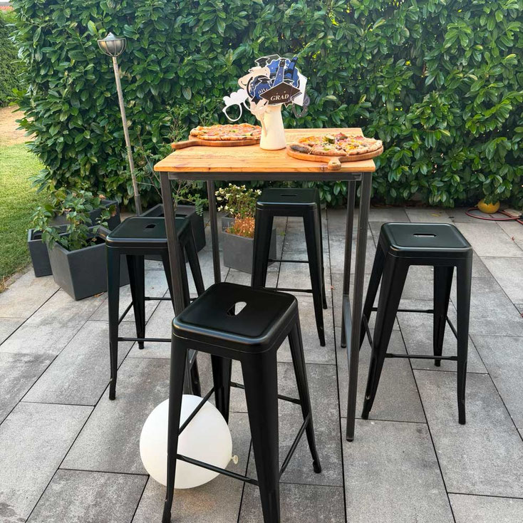 Stehtisch im industriellen Design mit dunkler Holzplatte. Dunkelgraues Tischgestell. Auf dem Tisch sind zwei Pizzas auf Holztellern serviert und eine Vase in der Mitte. Passende dunkle Stühle. Alle möbel stehen auf einem Steinboden.