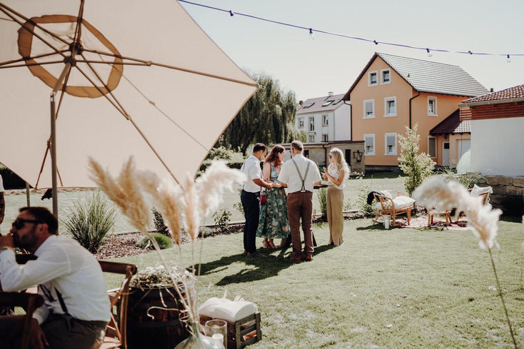 Sommerliche Gartenhochzeit bei Schweinfurt, die Gäste sind im Gespräch, großer Sonnenschirm, Lichterkette spannt sich über die Wiese, stilvolle Lounge-Möbel im Hintergrund zur Entspannung mit natürlicher Boho-Dekoration.