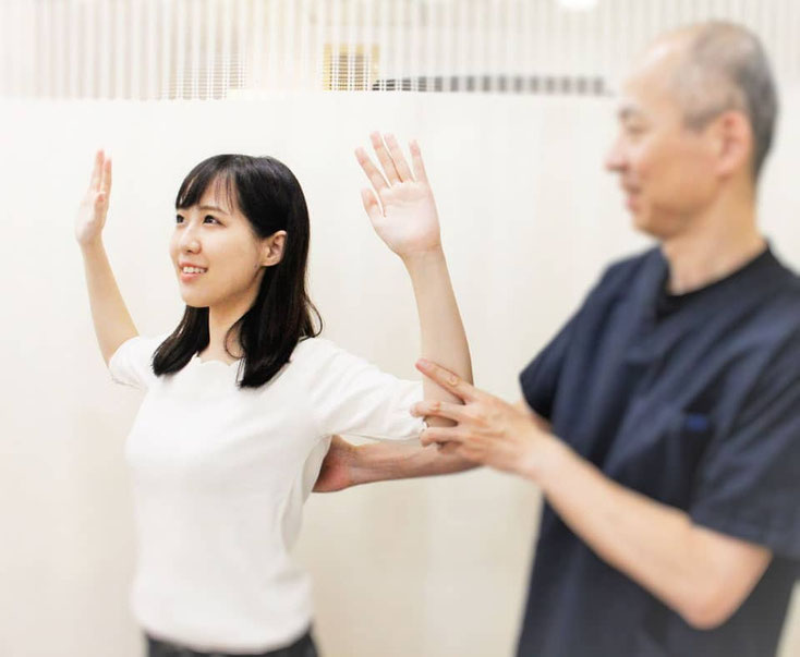 患者の女性が姿勢を正して両腕を「W」の字のように曲げて上げ、背中のストレッチをしています。男性院長が横から肘を支えて正しいフォームを指導しています。