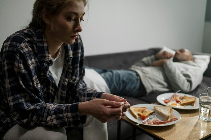 Foto: Eine Frau sitzt auf der Couch und sieht traurig zu Boden, dahinter liegt ein Mann auf der Couch, vor ihnen am Couchtisch unberührte Teiller mit Essen