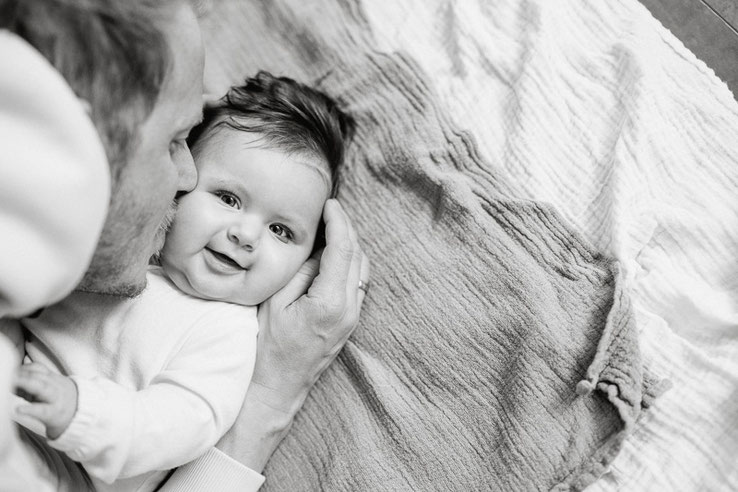 Ein fröhliches Baby wird im Liegen freudestrahlend vom Vater auf die Wange geküsst.