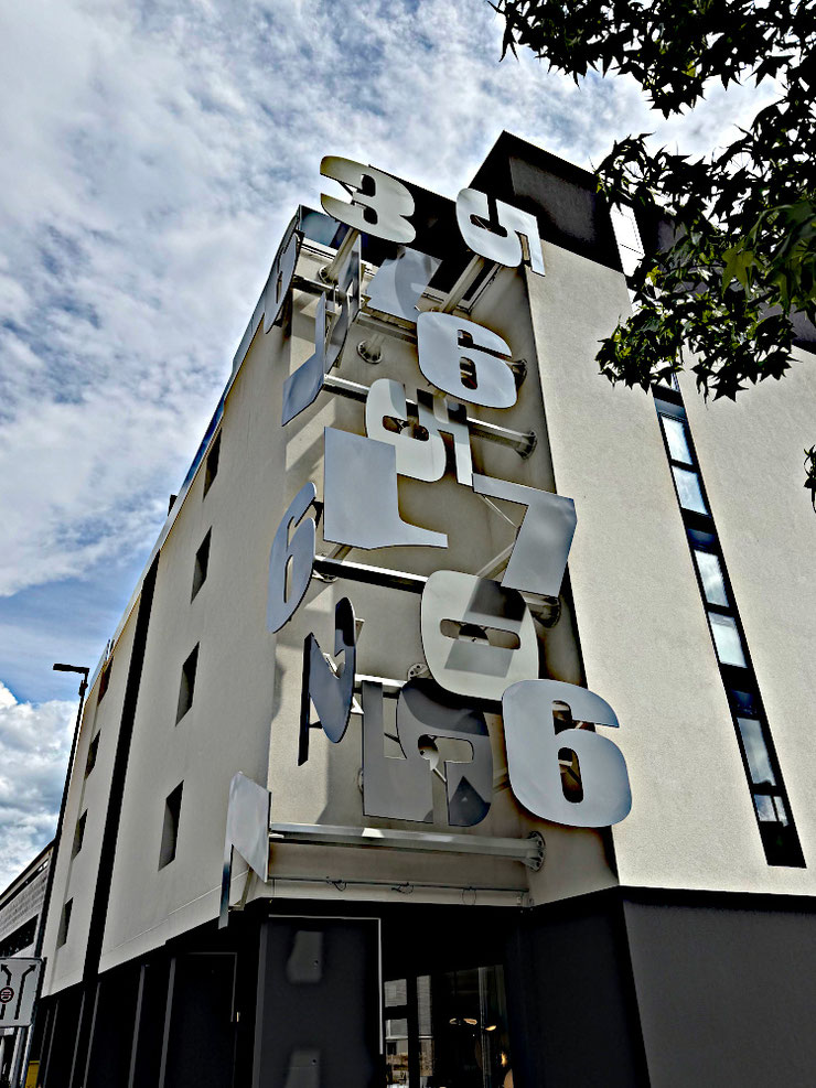 Street-level view of Paolo Grassi’s public installation in Massagno, showing oversized metallic numbers suspended along the building's vertical axis.