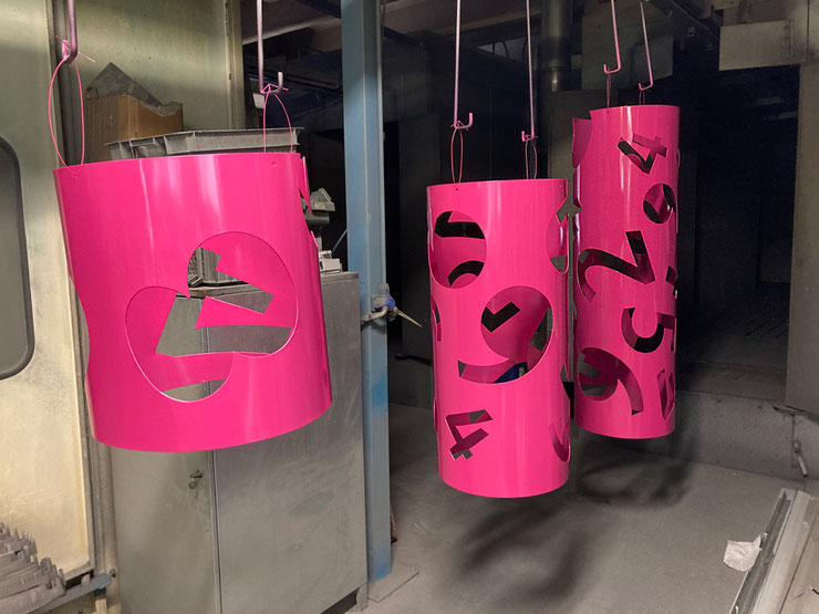 paolo-grassi-pink-panther-assembly-process – Bright pink sculptural cylinders with numeric cut-outs suspended in a workshop during the powder-coating phase.