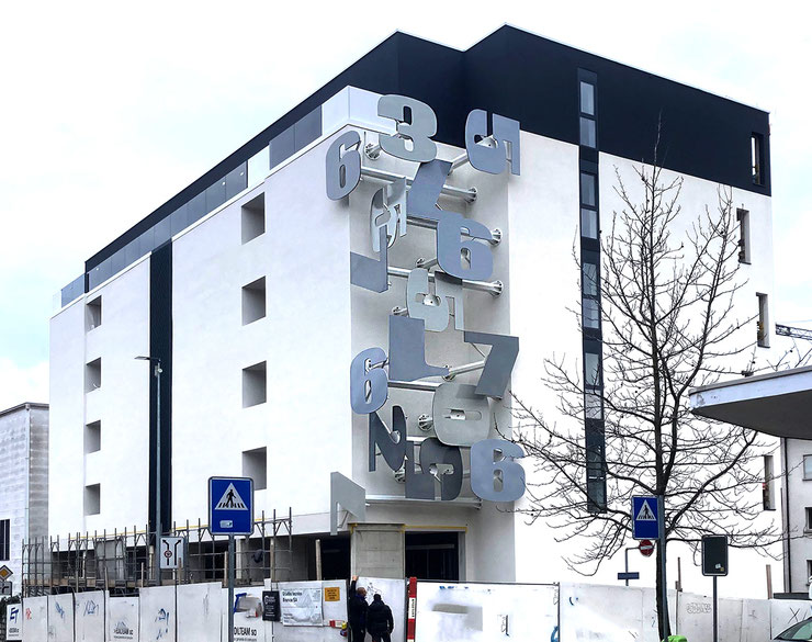 Conceptual public installation by Paolo Grassi in Massagno, featuring large metallic numbers emerging from a residential building facade.