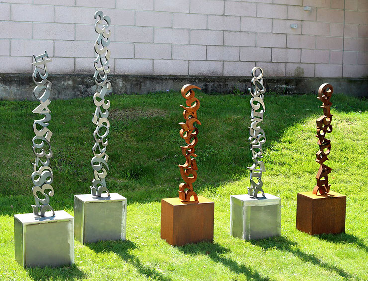 Selezione di cinque sculture verticali dalla serie Steli 2.0 di Paolo Grassi, con elementi alternati in acciaio inox e acciaio corten. Le opere indagano il ritmo dei numeri in una sequenza spaziale.