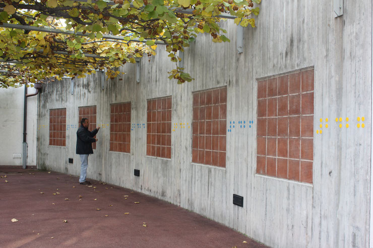 Side view of a long outdoor wall with colored Braille letters and a person interacting with the work