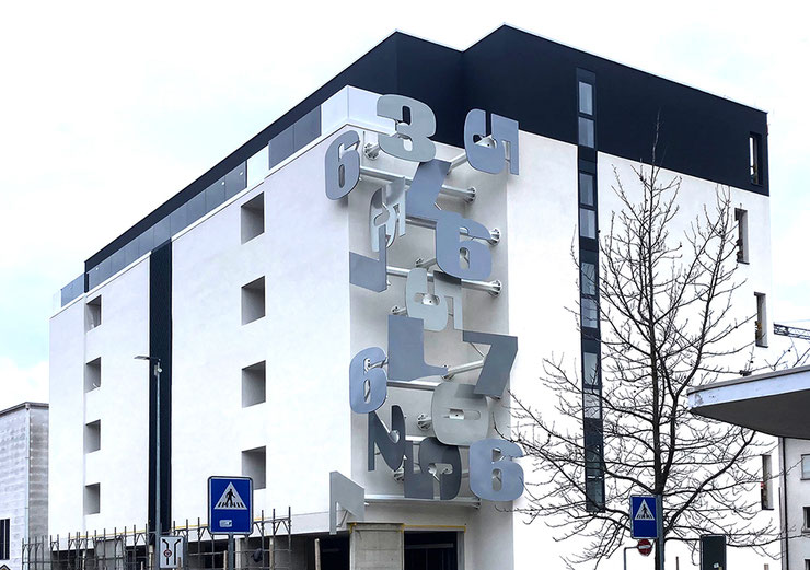 Site-specific conceptual installation by Paolo Grassi on a building corner in Massagno, featuring dynamic silver numbers projecting into urban space.