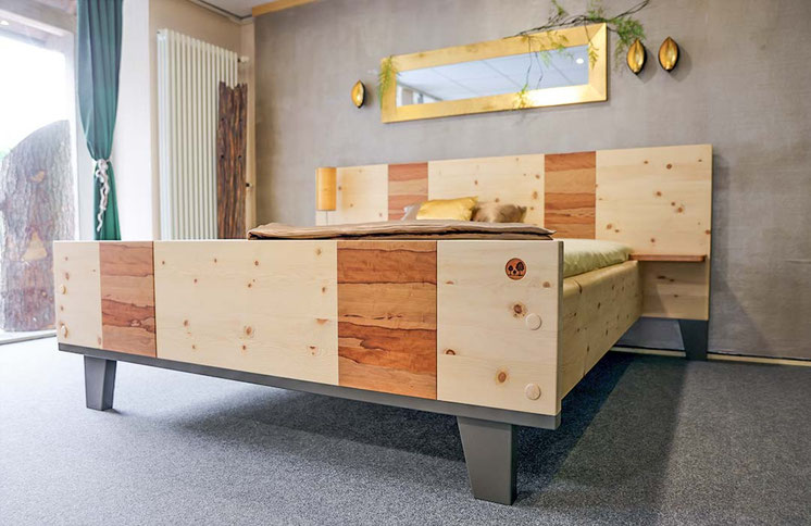 Modernes Zirbenbett „Goldbirne“ vor einer grauen Betonwand. Das Fußteil und das Kopfteil haben jeweils zwei Paneele aus rötlich-braunem Birnbaumholz, was einen kontrastreichen Look erzeugt. Das Bett steht auf vier mattschwarzen, konischen Füßen
