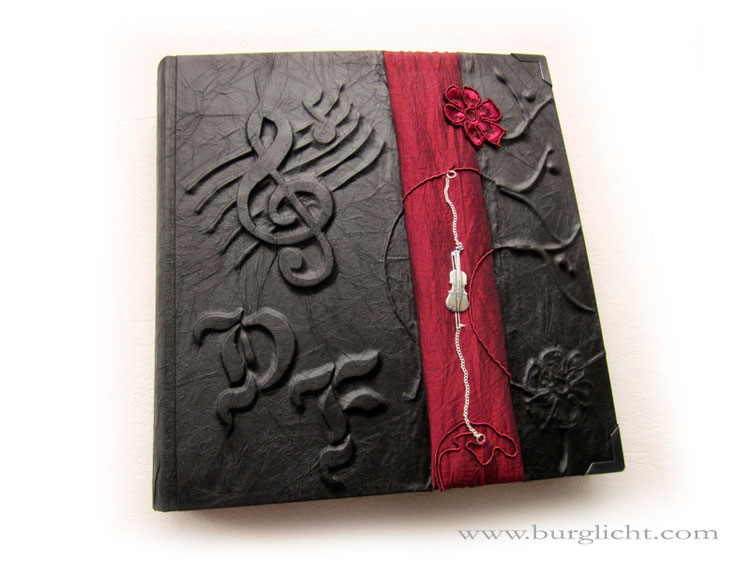 Fotoalbum - 29cmx32cm, 100 Seiten schwarz, Bezug: Stoff schwarz, Hochrelief Initialen Notenschlüssel Ranken Blume, vertikale Borte Taft rot mit Anhänger Geige.