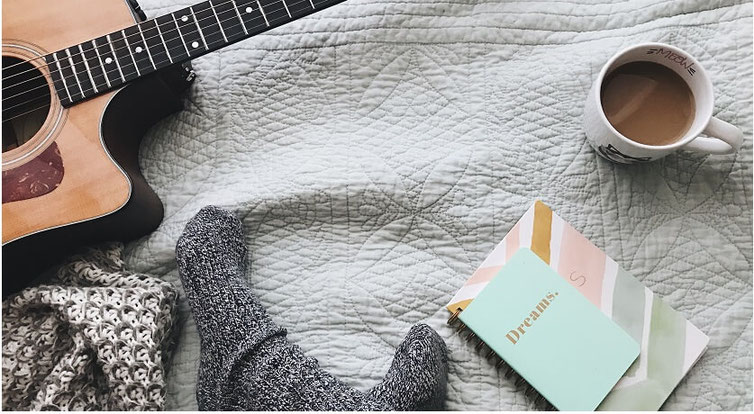 Feet in wool socks on a bed with a guitar, cup of coffee, journal and cozy-looking blanket