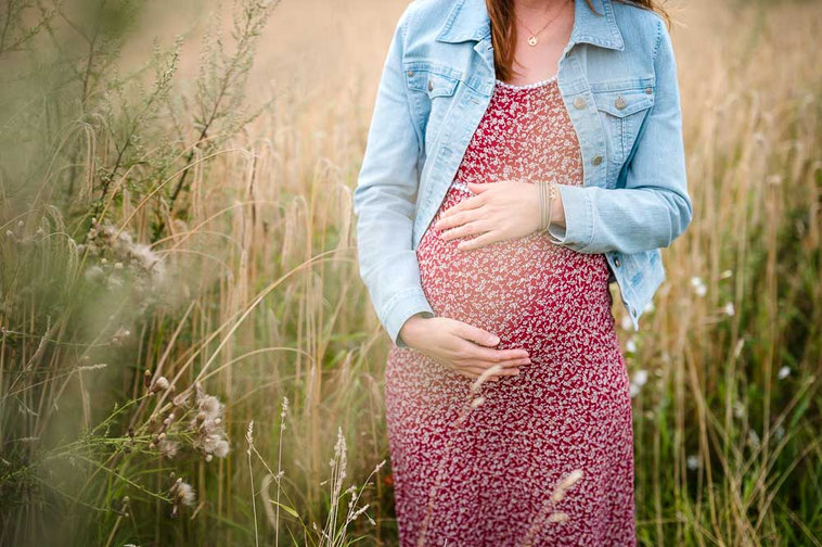 Eine Schwangere steht im roten Kleid und Jeans-Jacke auf einer sommerlichen Wiese und hält dabei ihren Babybauch fest.
