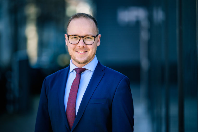 Lächelnder junger Mann mit Brille, Krawatte und blauem Anzug in einer Tageslicht-Aufnahme.
