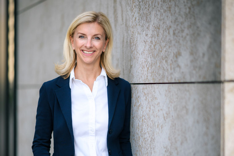 Lächelnde Frau im blauen Blazer und weißer Bluse, die entspannt an einer Wand lehnt.