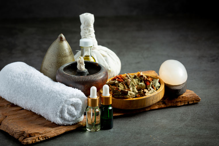 Spa-Utensilien wie ein weißes Handtuch, Ölfläschchen, Kräuterstempel, getrocknete Kräuter und ein glatter Stein auf einem Holzbrett.