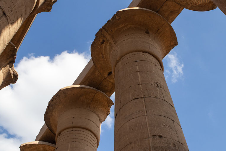 Massive ancient Egyptian stone columns with hieroglyphs, viewed from below against a bright blue sky with scattered clouds.