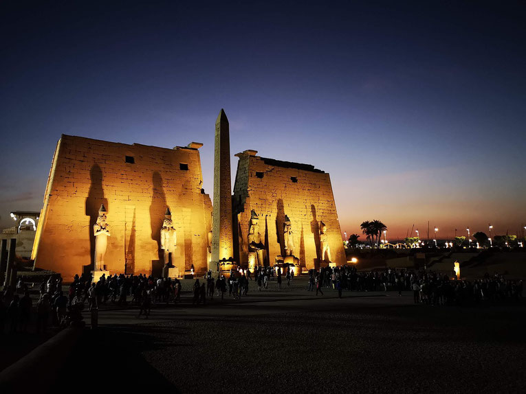 Illuminated statues and obelisk at the entrance of Luxor Temple, with crowds gathered at twilight under a fading sunset sky.