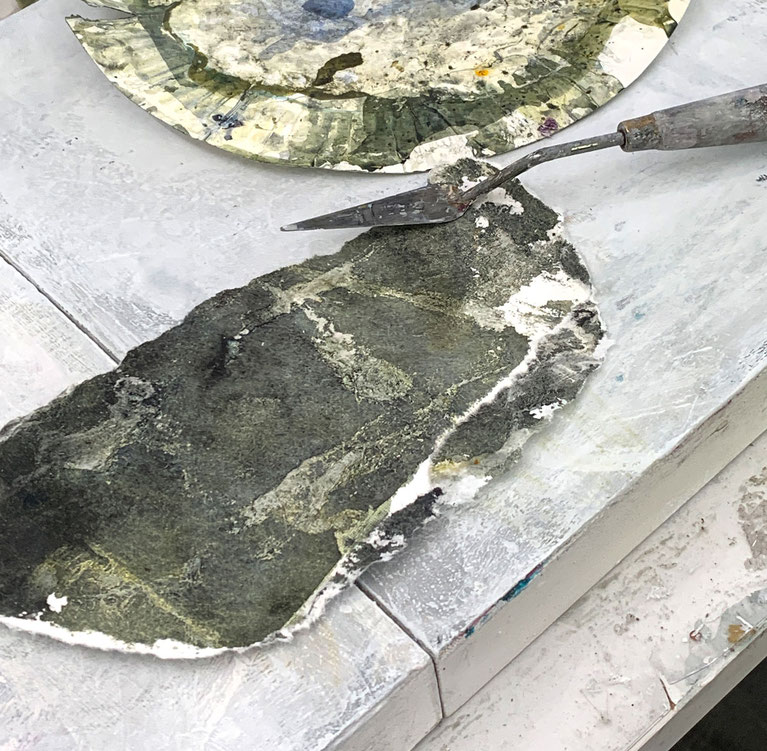Ein alter Pappteller mit Farbe, eine Malerspachtel und ein plastisch geformtes Papierfragment in einer Schüssel im Atelier.