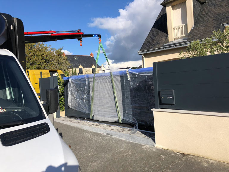 Une grue décharge un grand objet emballé et protégé par une bâche, devant une maison. L'objet est soulevé à l'aide de sangles depuis un camion.