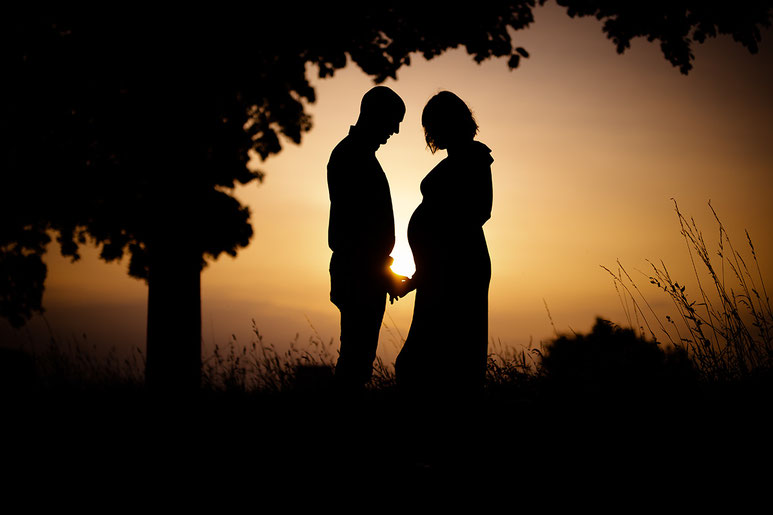 Schwangere Frau und Partner stehen sich bei Sonnenuntergang gegenüber und halten Hände.