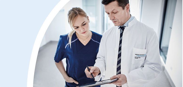 Ein Arzt und eine Krankenschwester in medizinischer Berufskleidung besprechen gemeinsam Informationen auf einem Tablet in einem Krankenhausflur.