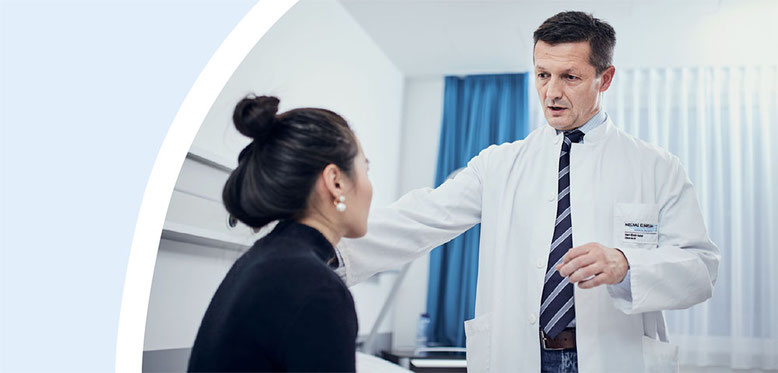 Ein Arzt in einem weißen Kittel mit Namensschild und Krawatte lächelt und spricht mit einer auf einem Bett sitzenden Patientin in einer Klinik.