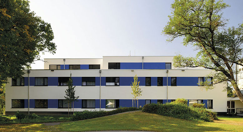 Außenansicht des Medias Klinikums Burghausen, modernes weiß-blaues Gebäude umgeben von Bäumen und Grünflächen.