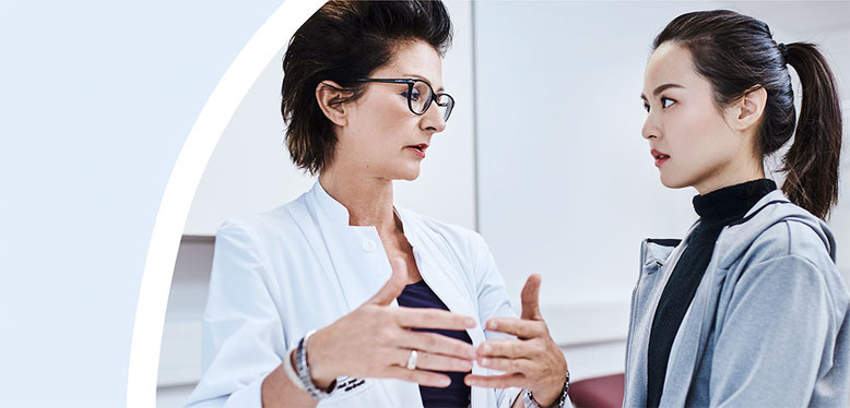 Ärztin im weißen Kittel spricht ernsthaft und erklärend mit einer Patientin in einem modernen Behandlungszimmer.