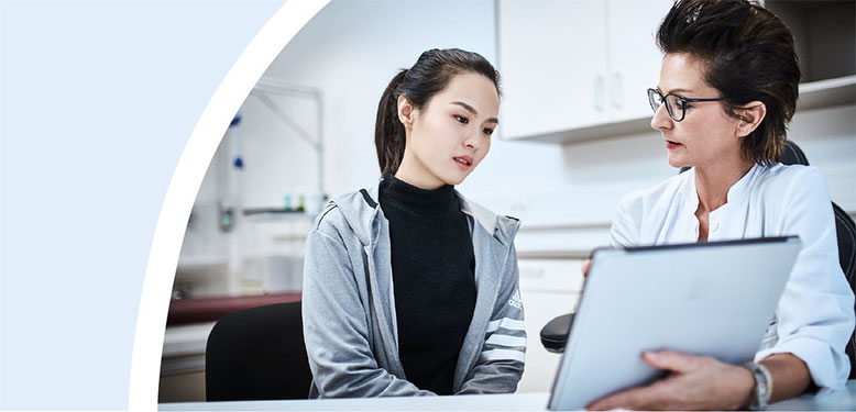 Ärztin in weißem Kittel zeigt einer Patientin Informationen auf einem Tablet.