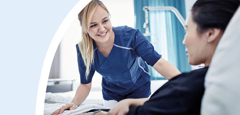 Lächelnde Krankenschwester in blauer Uniform, die sich im Krankenzimmer zu einer Patientin hinunterbeugt, die in einem Bett liegt.