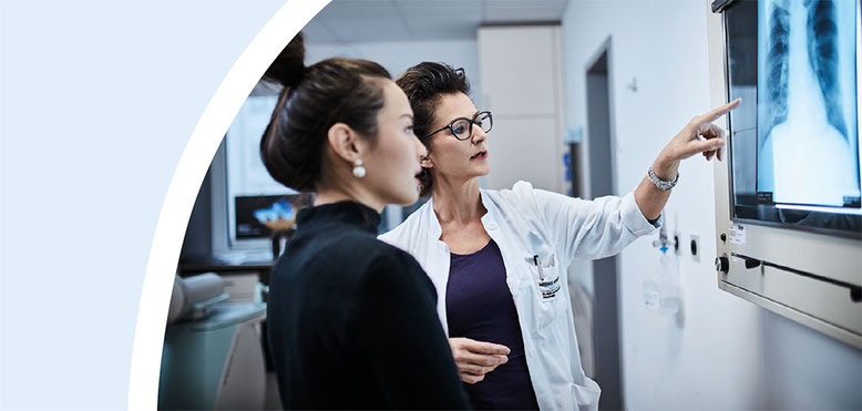 Eine Ärztin und ihre Patientin sind im Gespräch und blicken auf einen Leuchtkasten im Krankenhaus, in dem Röntgenaufnahmen gezeigt werden.