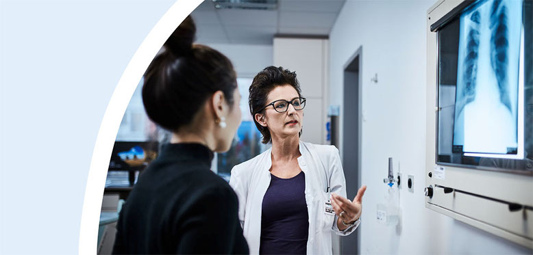 Ärztin im weißen Kittel erklärt einer Patientin ein Röntgenbild in einem modernen Behandlungsraum im Krankenhaus.