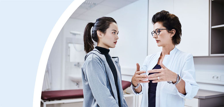 Ärztin im weißen Kittel spricht ernsthaft und erklärend mit einer Patientin in einem modernen Behandlungszimmer.