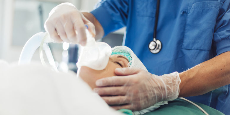 Arzt in blauer OP-Kleidung setzt einer Patientin mit einer Atemmaske vor der Narkose die Maske auf, um sie für die Operation vorzubereiten.