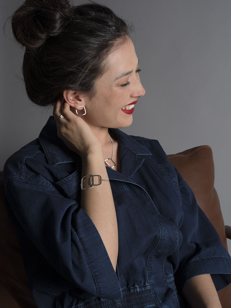 jeune femme souriante assise dans un fauteuil en cuir brun portant un bracelet Q06, un collier Q03 et des boucles d'oreilles Q08 en argent recyclé de la collection Quadra