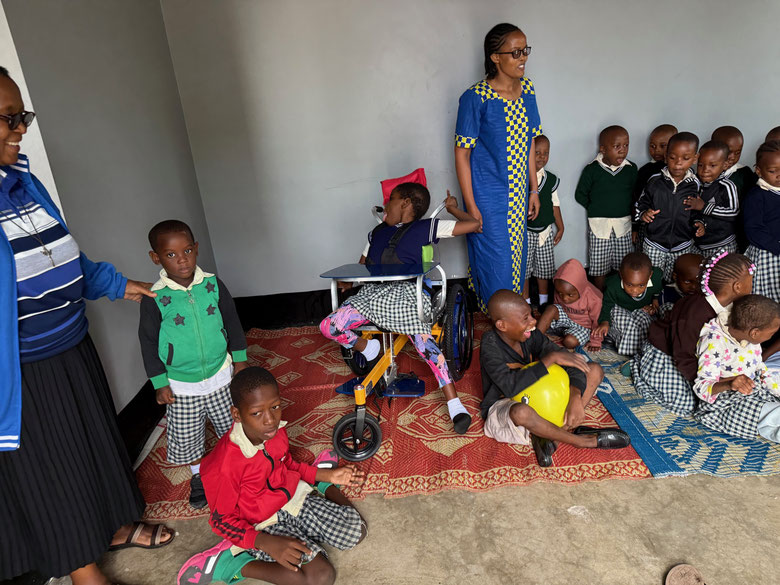 Betreuerin steht hinter einer Gruppe behinderter Kinder, teils im Rollstuhl oder auf dem Boden sitzend, im Klassenraum