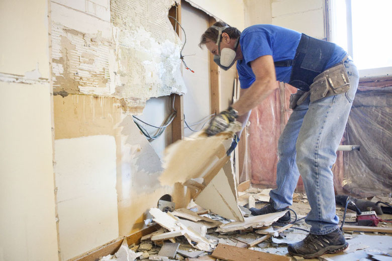 The kitchen demolition is in full swing, with hammers pounding, tiles shattering, and countertops being pried loose amidst a cloud of dust and debris.