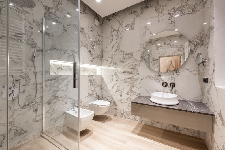 This is a modern bathroom featuring a sleek design with marble walls, a walk-in glass shower, a floating vanity with a round mirror, and both a wall-mounted toilet and bidet, complemented by warm wood-effect flooring.